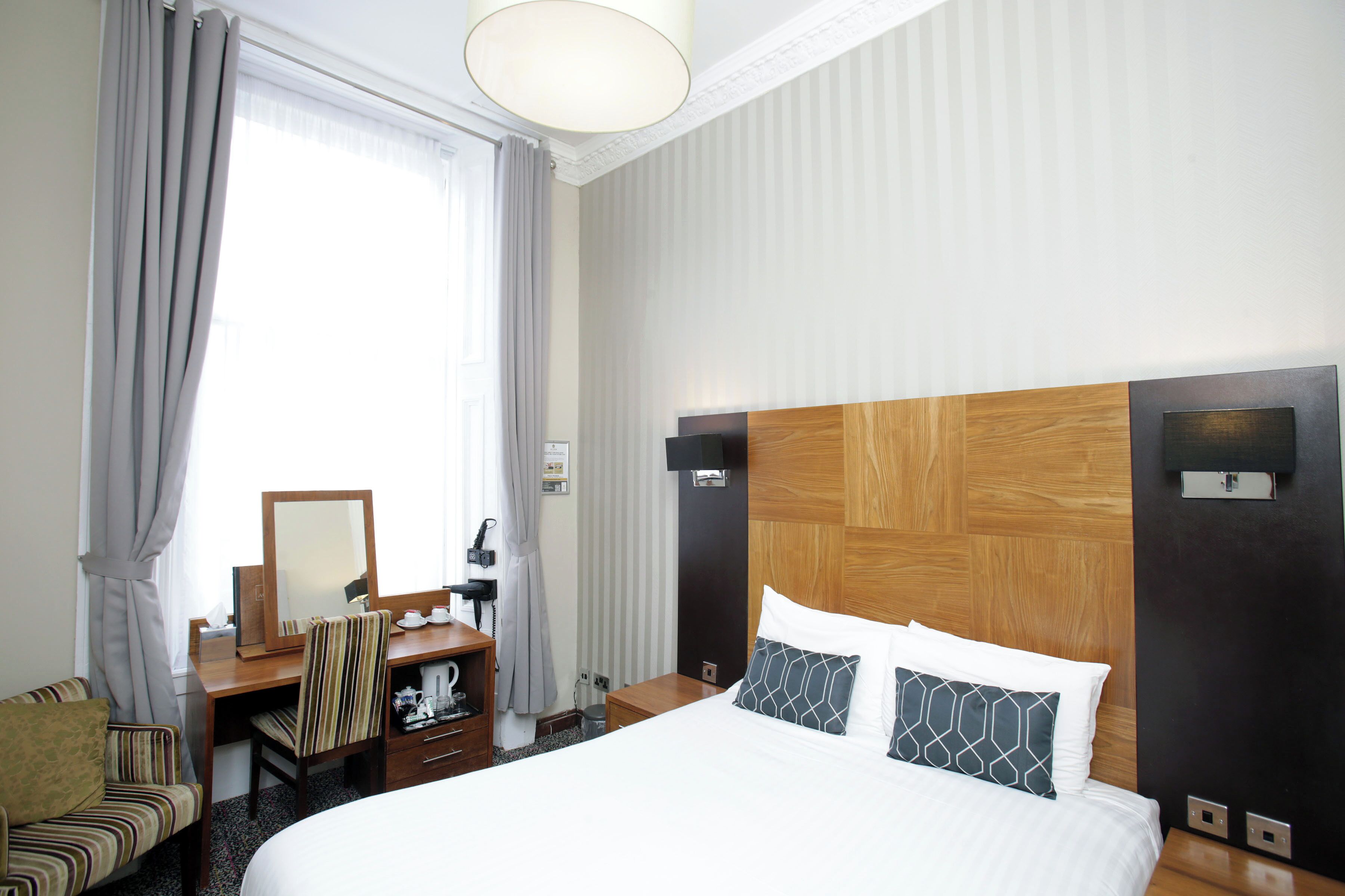 Une chambre d’hôtel avec un grand lit, une tête de lit en bois, un bureau avec un miroir, une chaise et une fenêtre avec des rideaux.