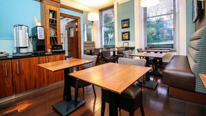 A restaurant with wooden tables and chairs, a counter with a coffee machine, and a large window with blinds. - Acorn Hotel (Glasgow)
