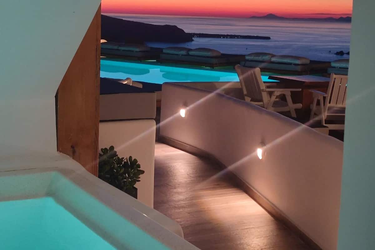 A balcony with a view of the ocean at sunset, a hot tub, and outdoor seating.