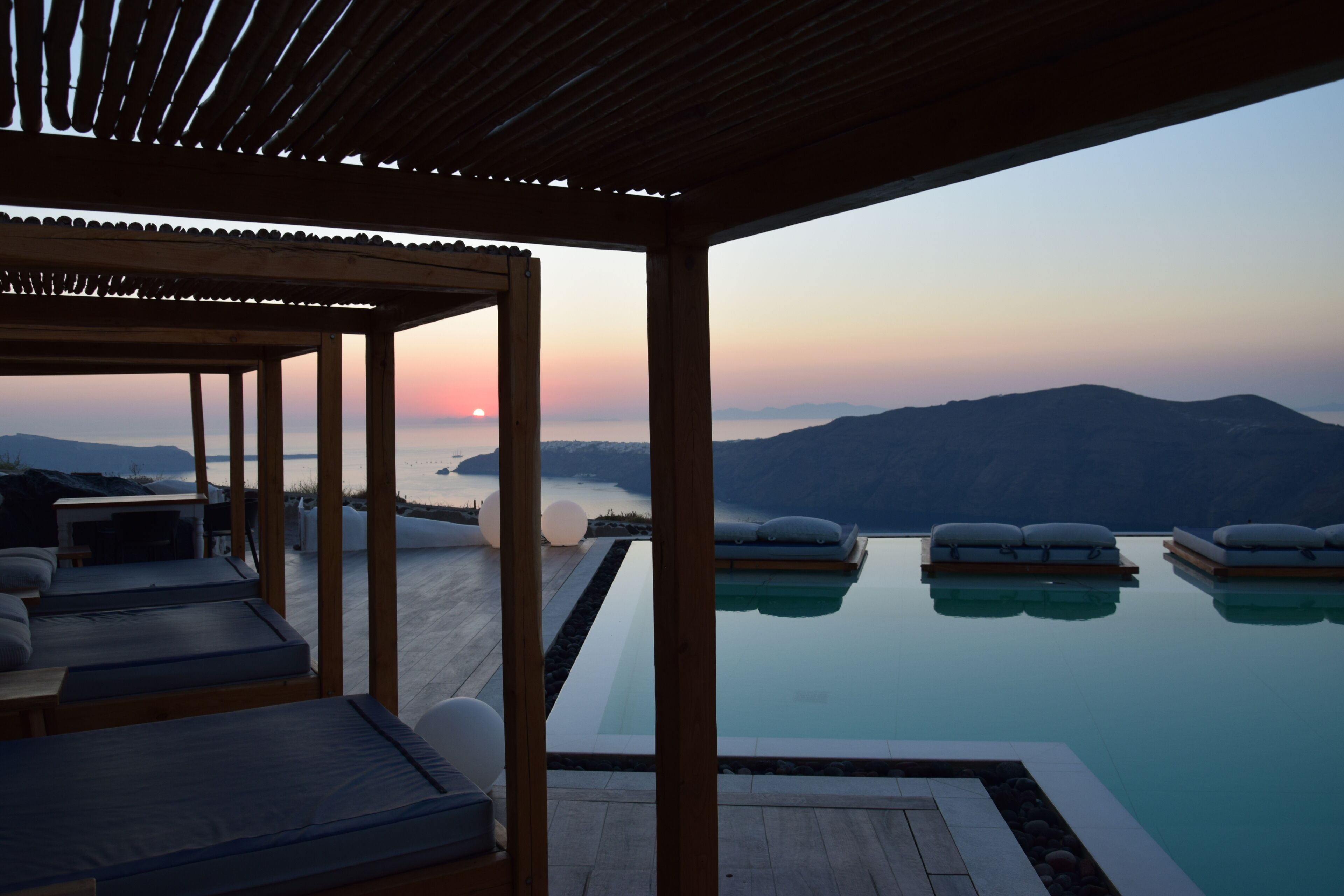Uma piscina infinita com vista para o pôr do sol, área de estar com deck de madeira e espaço coberto com almofadas.