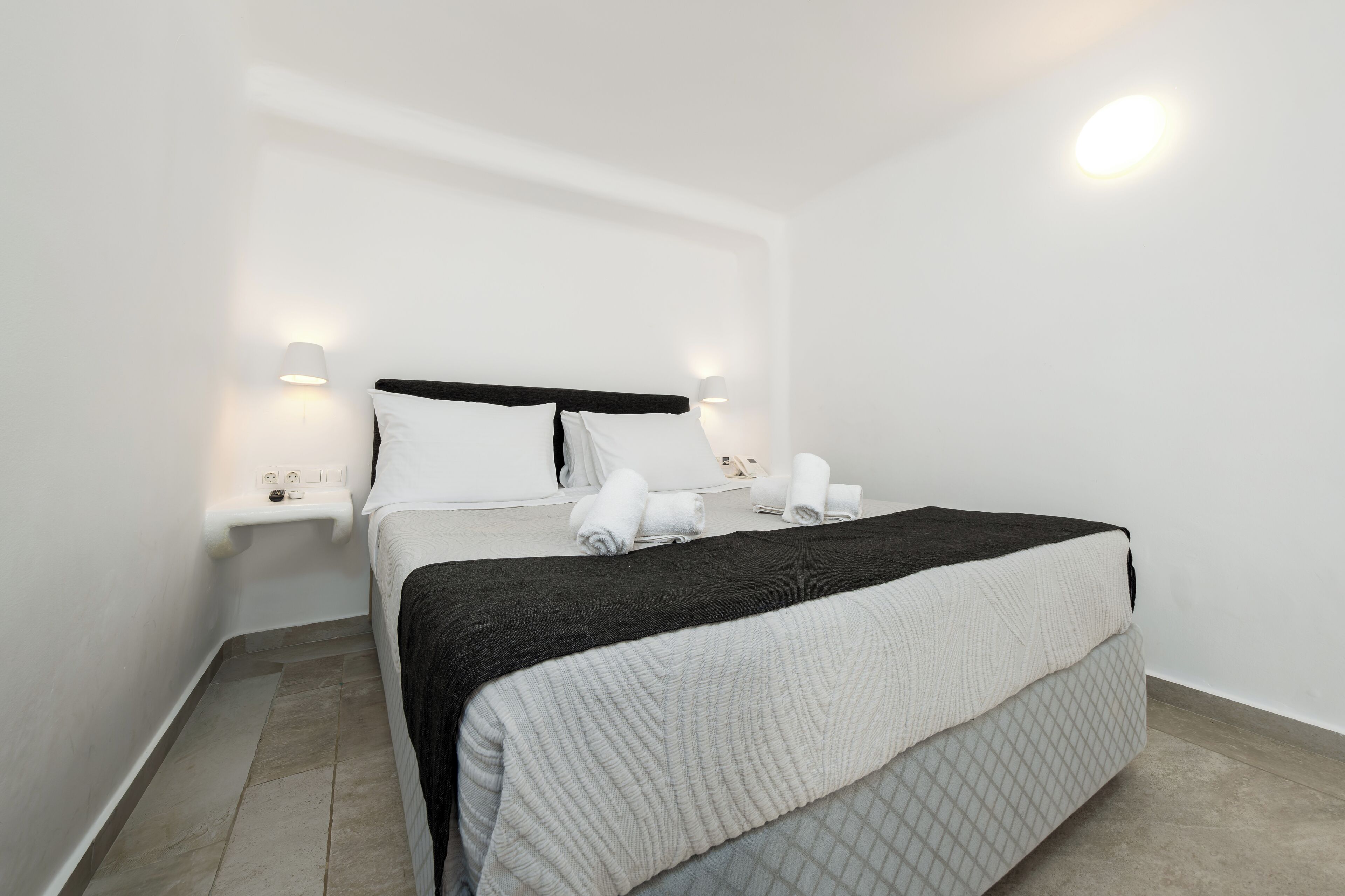 A neatly made bed with a black and white blanket, two pillows, and rolled towels on a white wall with a night light.