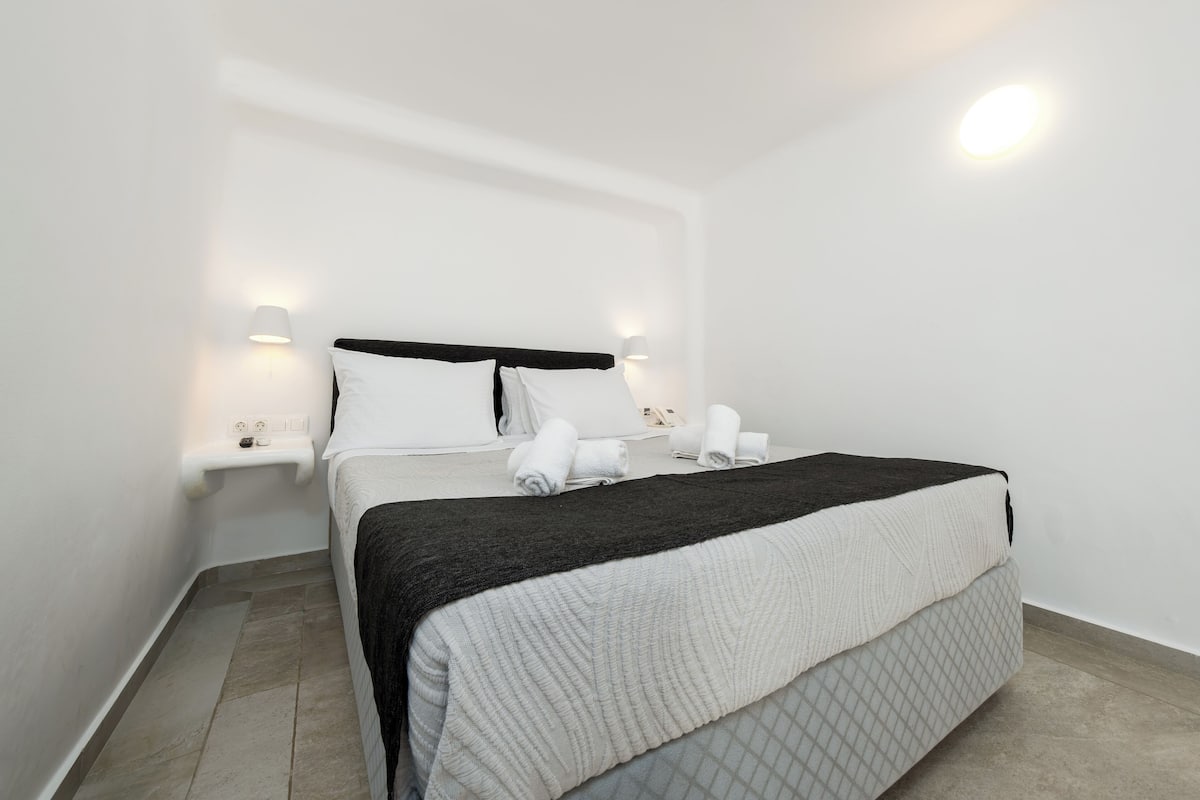 A neatly made bed with a black headboard, white pillows, and a white duvet. There are two bedside tables with lamps and a small shelf with folded towels.