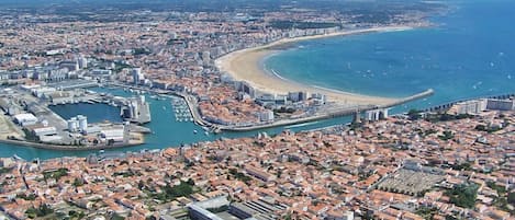 Vista aérea de uma cidade costeira com um porto de iates, uma praia e áreas urbanas densas.