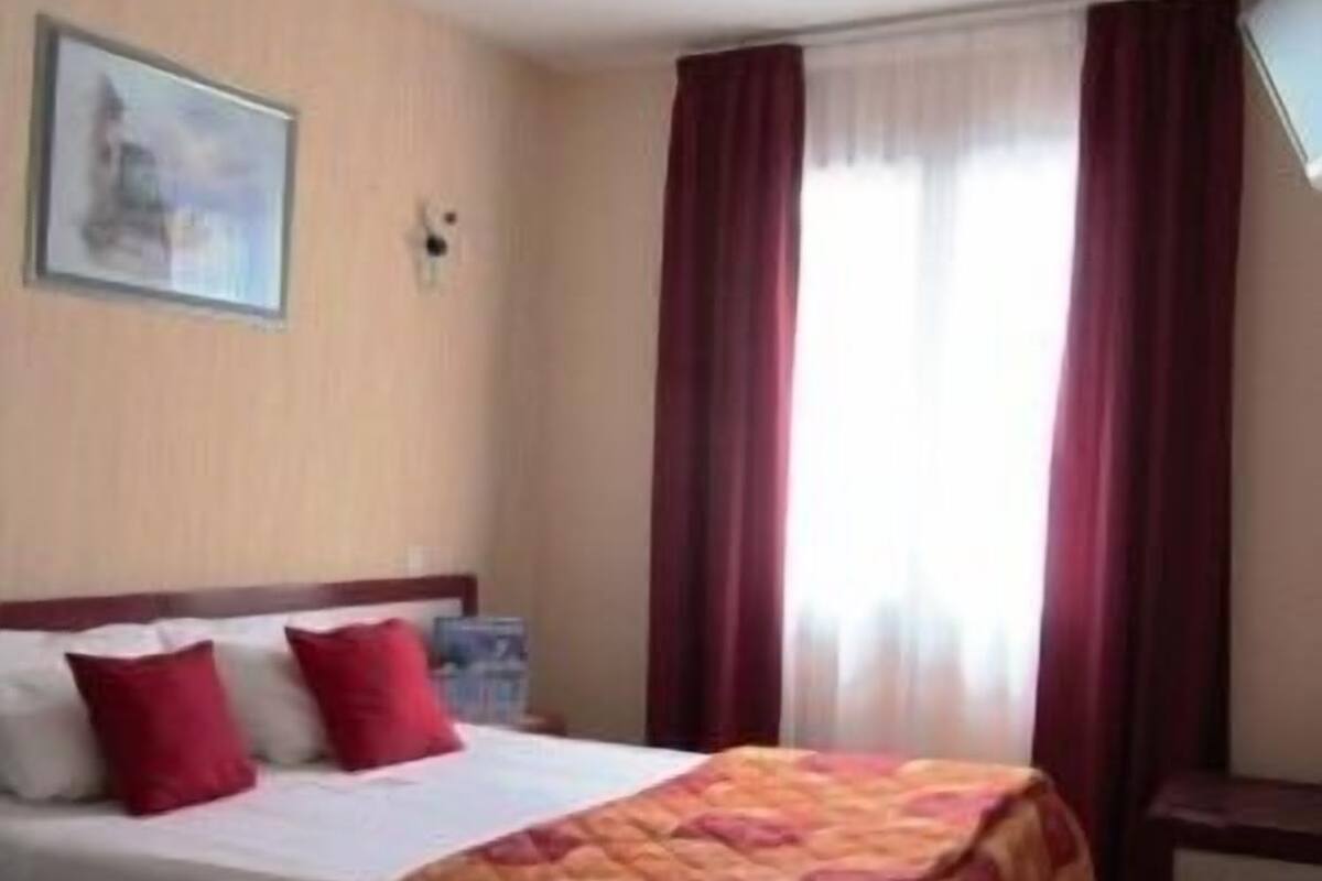 Habitación de hotel con cama, cortinas rojas, un cuadro en la pared y una ventana con cortinas de encaje.
