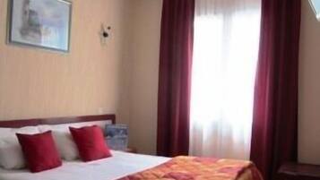 A hotel room with a bed, red curtains, a painting on the wall, and a window with sheer curtains.