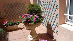 Una terraza con geranios en macetas, un cerramiento de celosía y un edificio con techo de tejas.