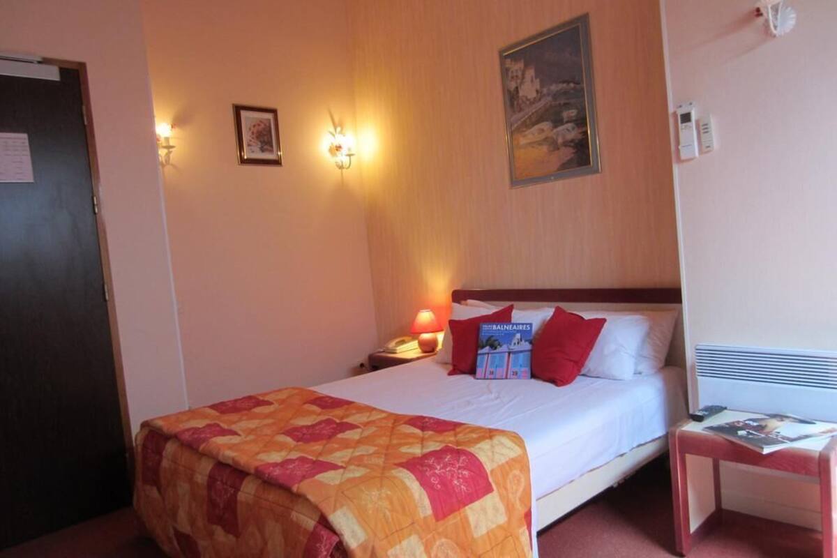 Habitación de hotel con cama, mesita de noche, lámpara, un cuadro enmarcado y una lámpara fijada a la pared.