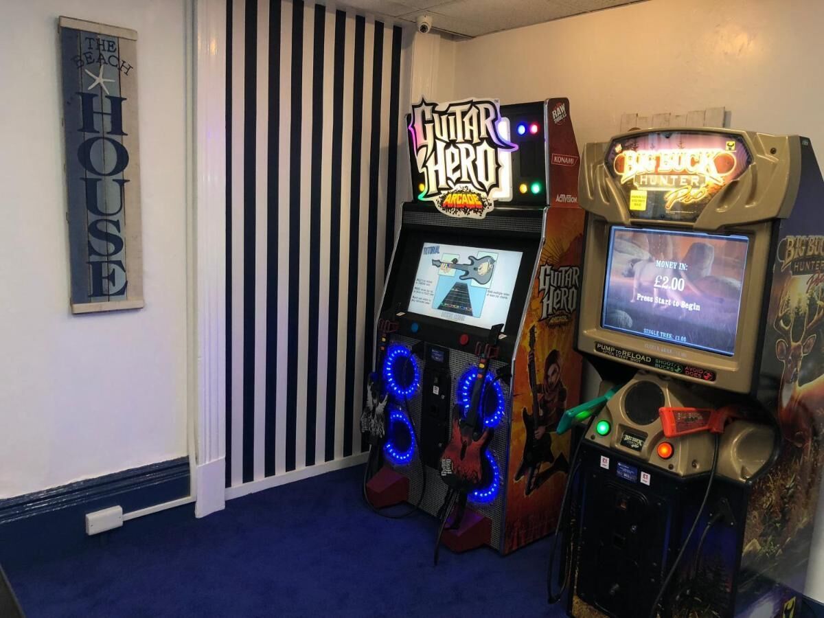 A room with a striped wall, a neon sign for 'Guitar Hero', and a 'Big Buck Hunter' game machine.