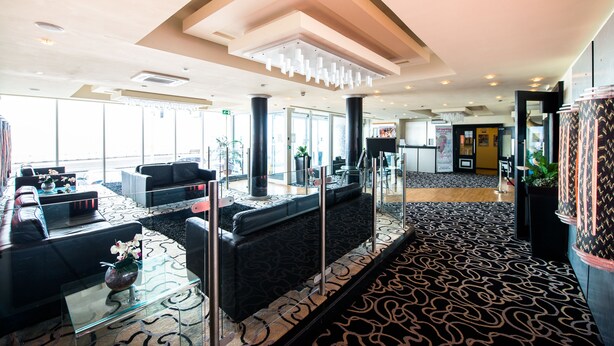 Un hall d’entrée spacieux au design moderne, avec un tapis à motifs noir et blanc, des canapés en cuir noir et une table basse en verre surmontée d’un vase.