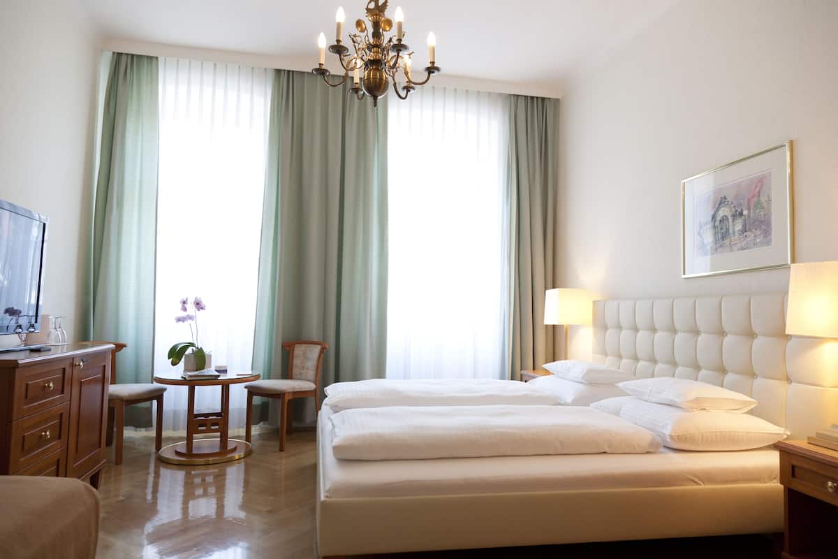 A hotel room with a large bed, a desk with a chair, a small table with a plant, and a chandelier.