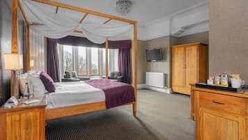 A four-poster bed with a canopy, a flat-screen TV mounted on the wall, a wooden wardrobe, and a bedside table with a lamp and a glass of water.