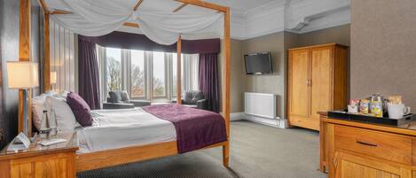 A four-poster bed with a canopy, a flat-screen TV mounted on the wall, a wooden wardrobe, and a bedside table with a lamp and a glass of water.