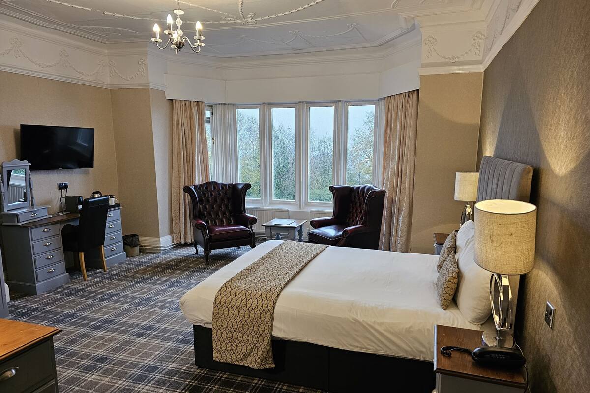 A hotel room with a large bed, two armchairs, a desk with a television, and a chandelier.