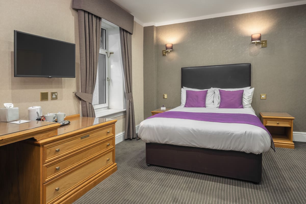A hotel room with a large bed, wooden dresser, bedside tables, a flat-screen TV, and a window with curtains.