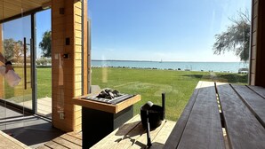 Una terraza de madera con una parrilla y una mesa, con vistas a una zona cubierta de césped y un cuerpo de agua con veleros.