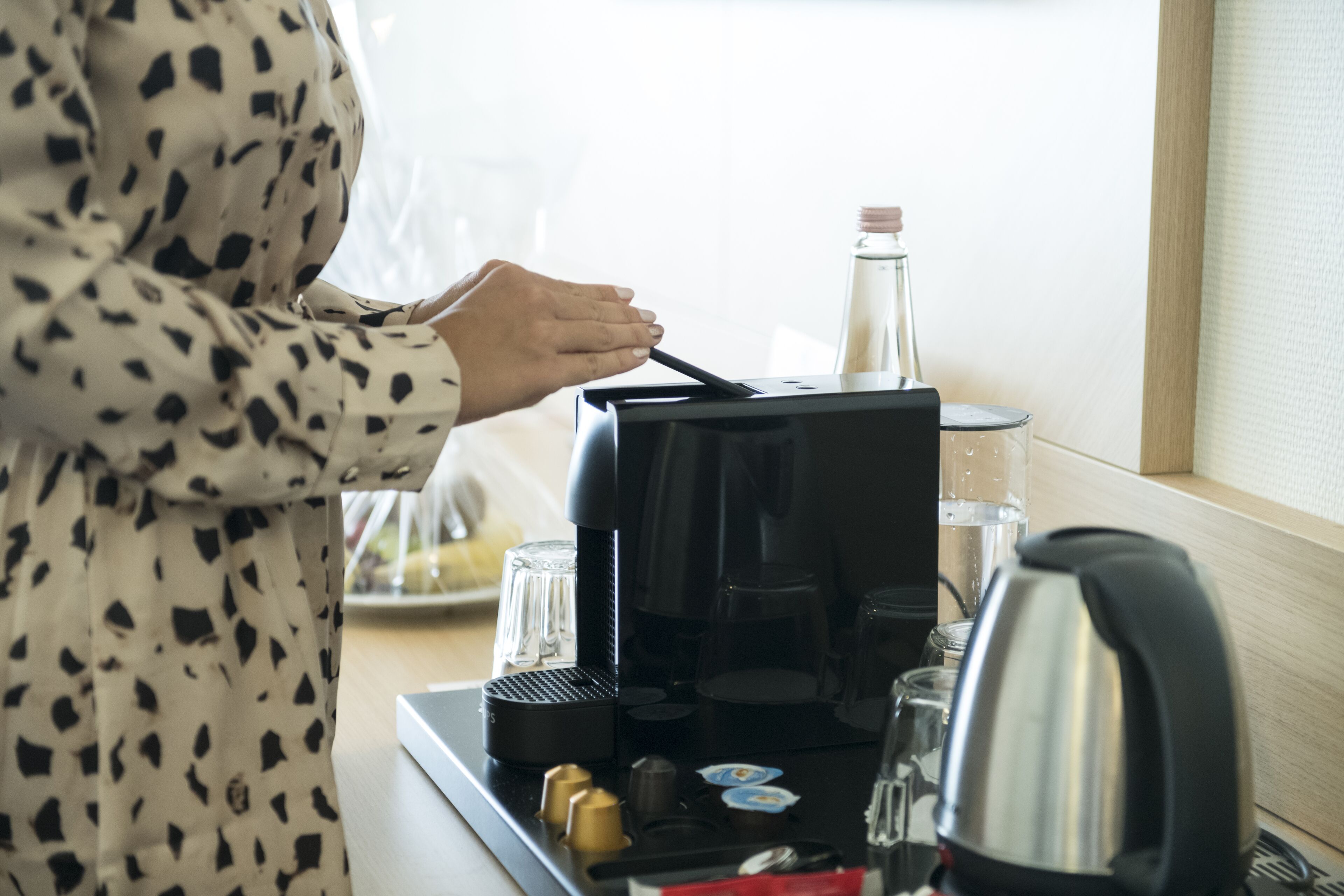 A person in a polka dot dress interacting with a coffee machine, with coffee cups and a kettle on the counter.