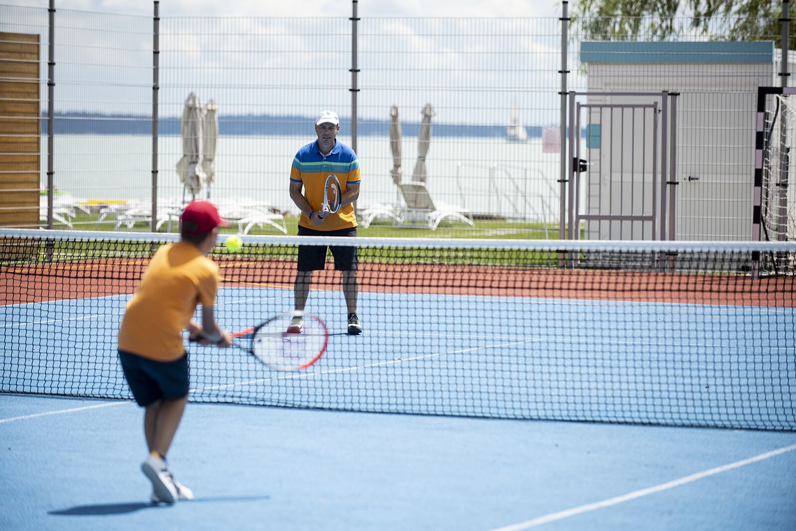 A tennis court with a net in the middle, a player in a yellow shirt and red cap, and another player in a blue and yellow shirt.