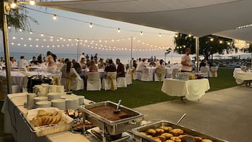 Evento al aire libre con mesas dispuestas bajo luces colgantes, bufé con pan y pasteles, e invitados cenando frente al agua.