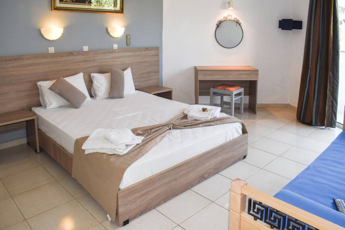A neatly made bed with a wooden headboard, a bedside table, a small desk with a chair, and a wall-mounted mirror.