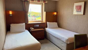 Habitación de hotel con dos camas individuales, una mesita de noche de madera con teléfono, una pequeña mesa y una ventana con cortinas.