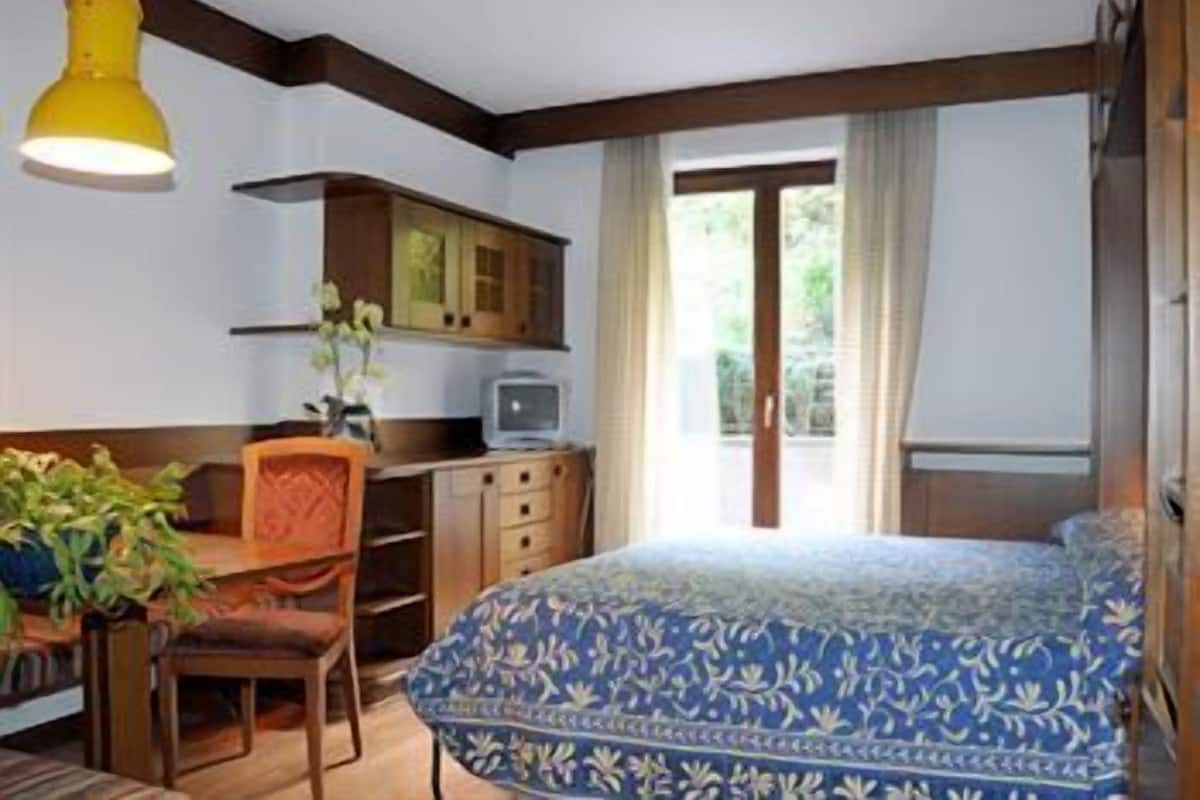 Un dormitorio con una cama, un escritorio con silla, un televisor y una ventana con cortinas.
