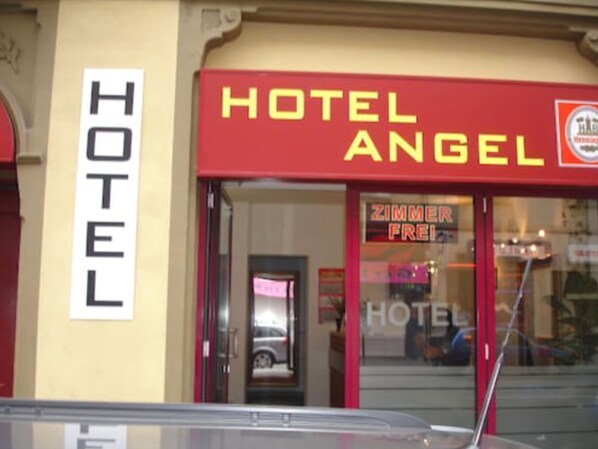 L’entrée d’un hôtel, avec une enseigne bien visible indiquant « HÔTEL ANGEL » et une plus petite enseigne indiquant « ZIMMER FREI » (chambre disponible).