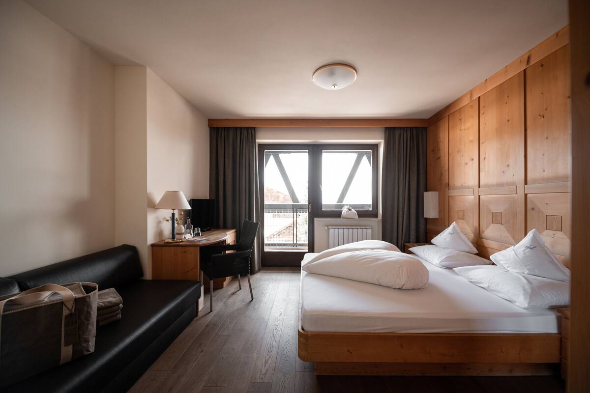 Habitación de hotel con una cama de madera, un sofá negro, un escritorio con una lámpara y vistas a través de un amplio ventanal.