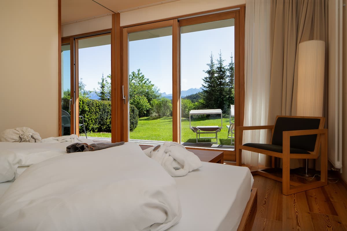 Habitación de hotel con una cama grande, una silla y vistas a un jardín y montañas a través de una puerta corrediza de vidrio.