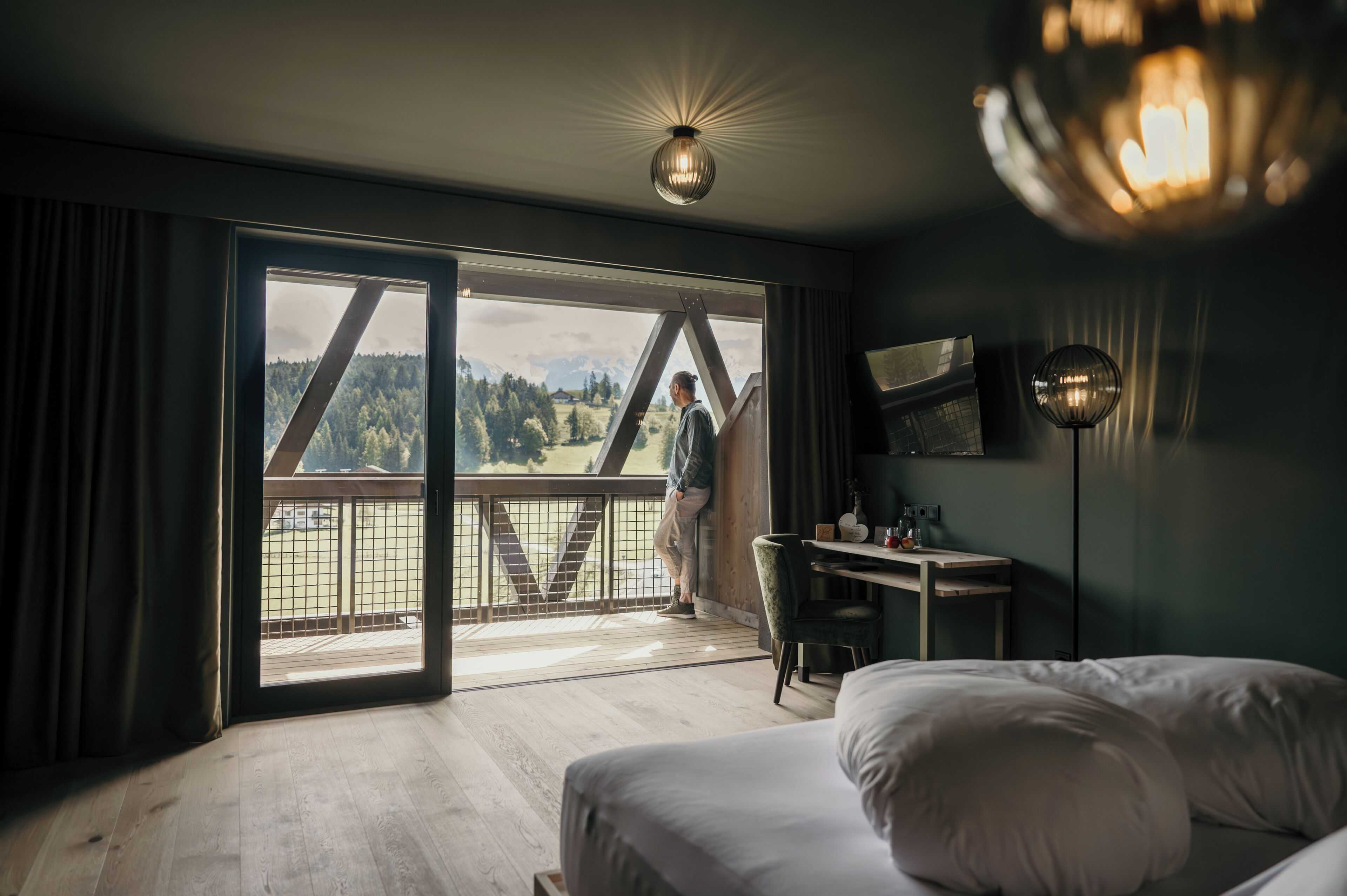 A hotel room with a large sliding glass door leading to a balcony, a bed, a desk, and a hanging light fixture.