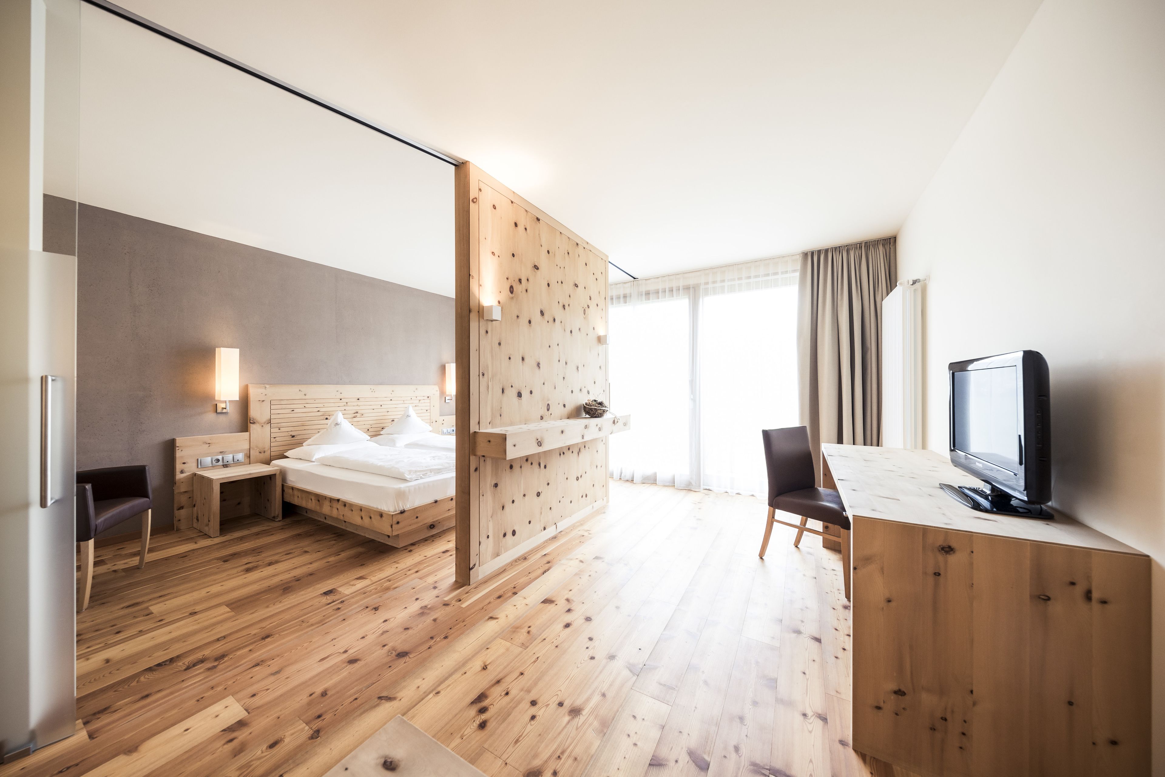 A modern hotel room with a wooden floor, a bed with white linens, a desk with a computer, a television, and a large window with curtains.