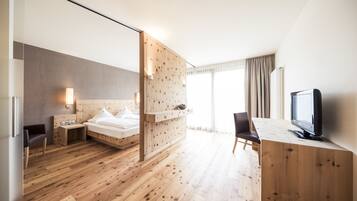 A modern hotel room with a wooden floor, a bed with white linens, a desk with a computer, a television, and a large window with curtains.