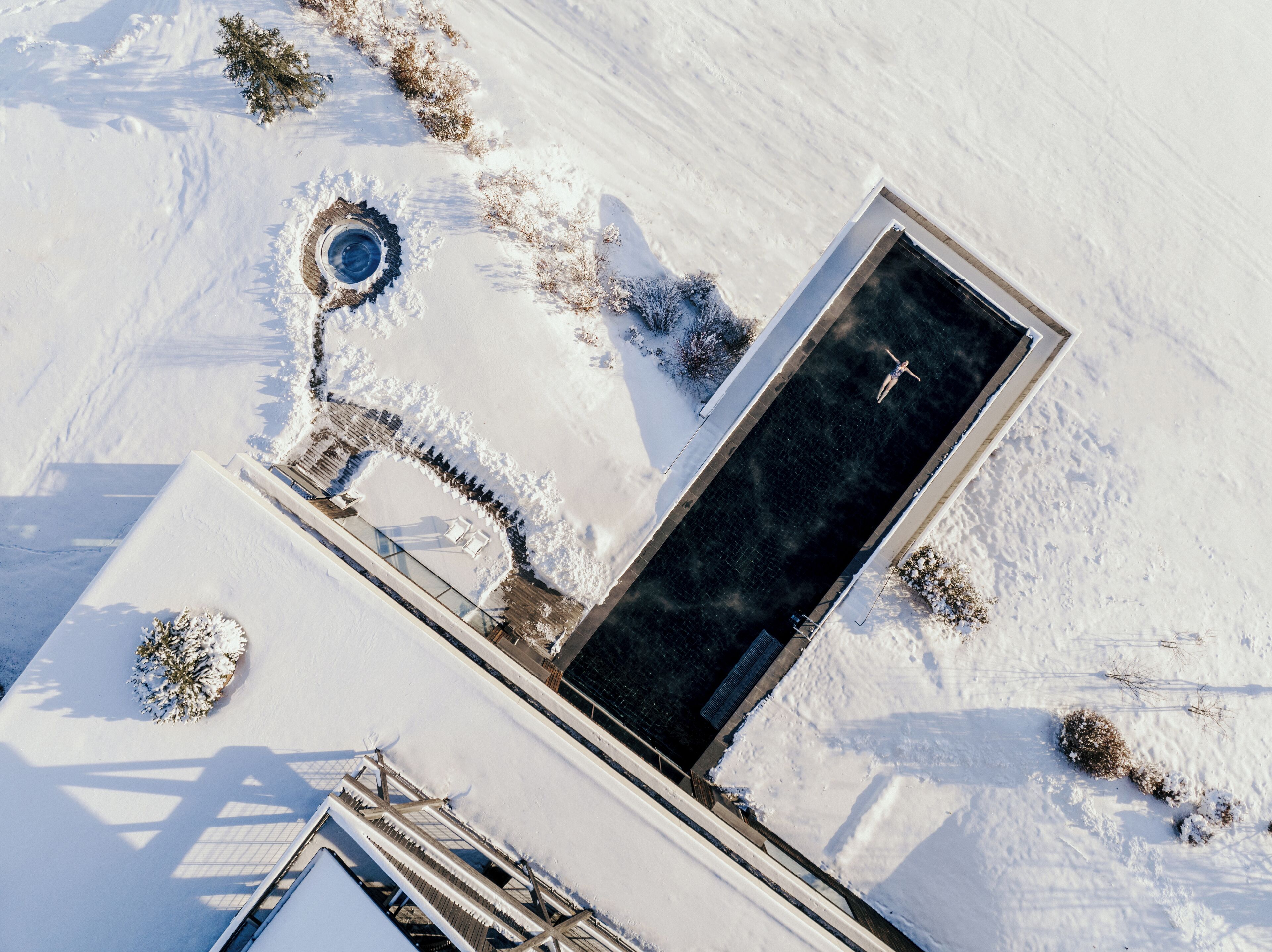 An aerial view of a snow-covered area with a rectangular pool, a small circular structure, and a few scattered bushes.