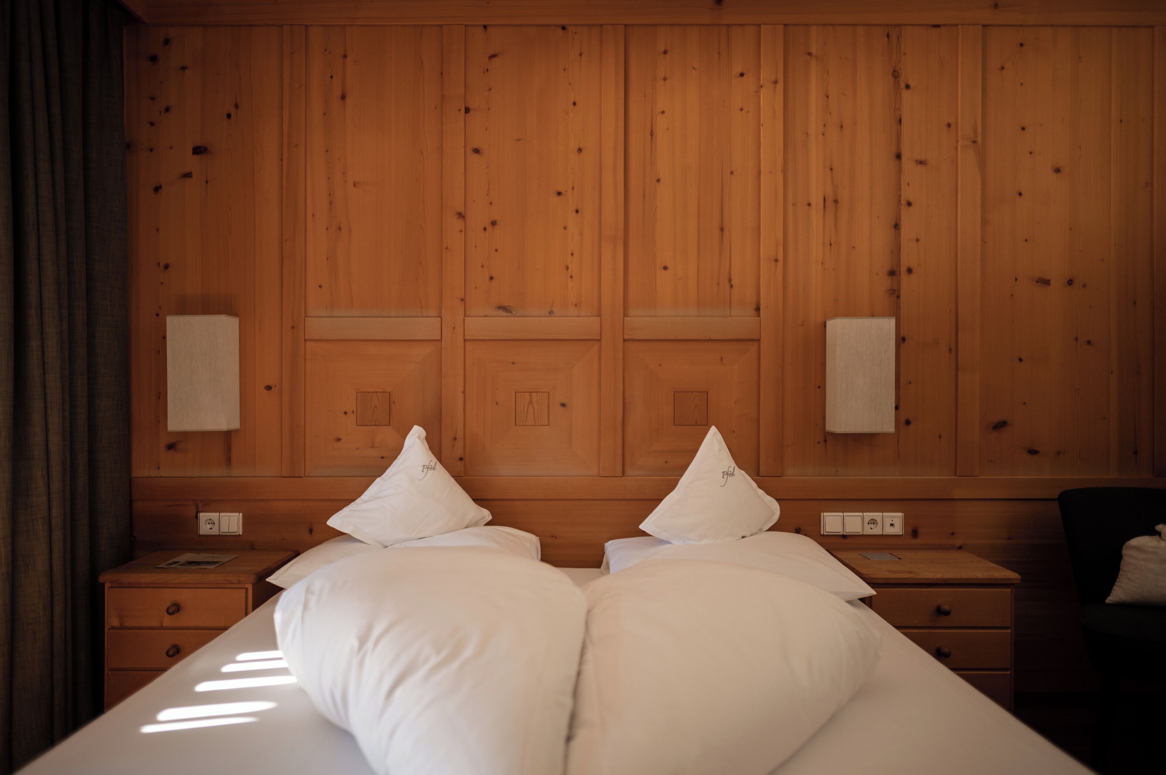 A hotel room with a wooden paneled headboard, two bedside tables, a bed with white bedding, and a chair.
