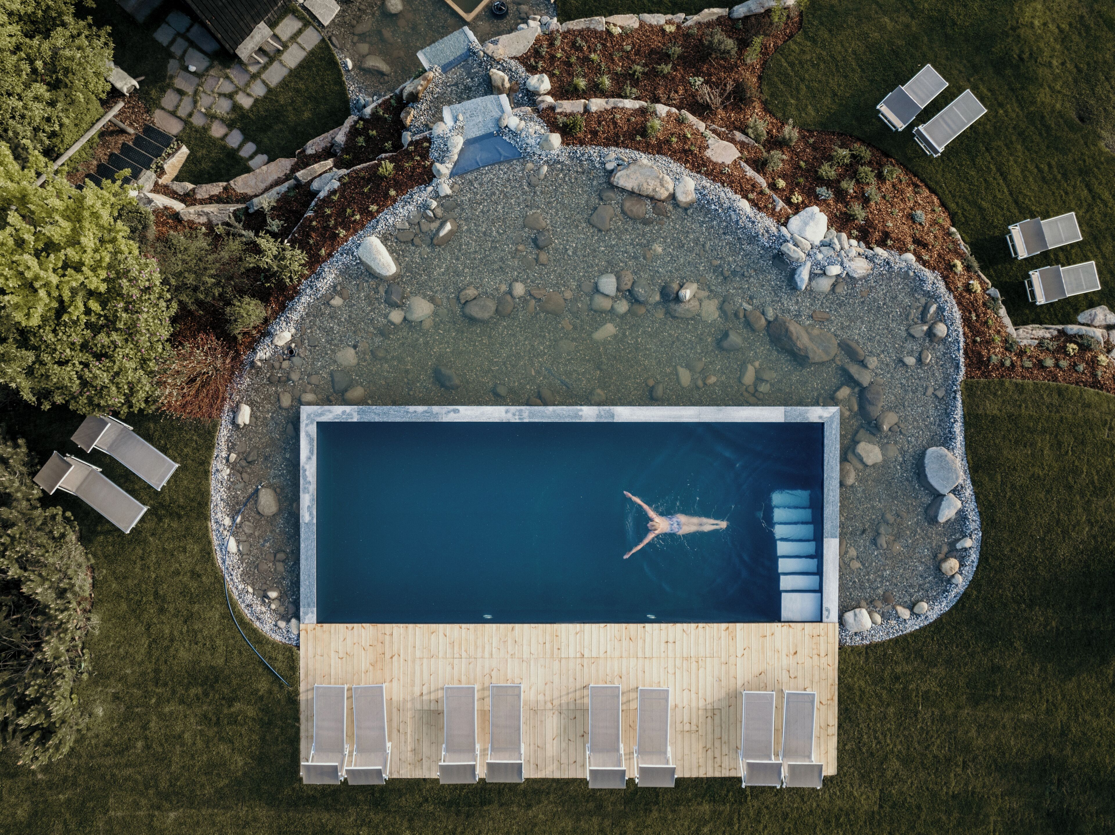 Eine Vogelperspektive auf einen Swimmingpool mit Holzterrasse, umgeben von Liegestühlen und einem angelegten Garten.