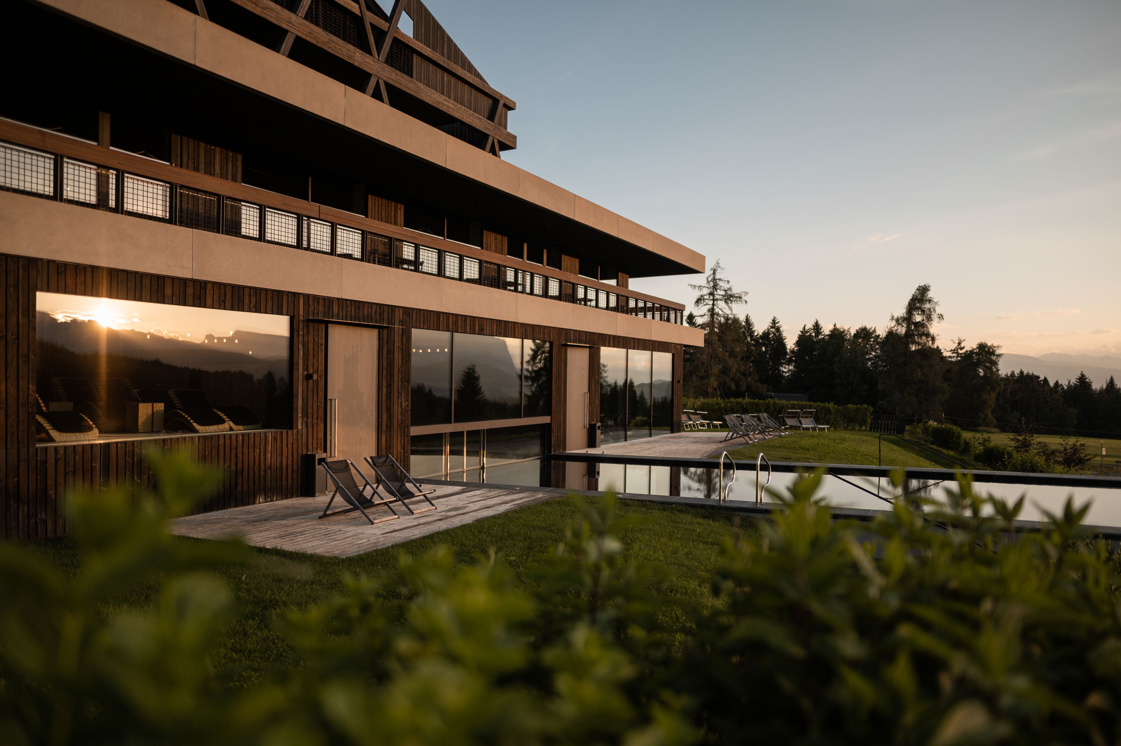 Un edificio moderno de dos pisos con amplios ventanales, una terraza con sillones de descanso y vistas a un lago y árboles.