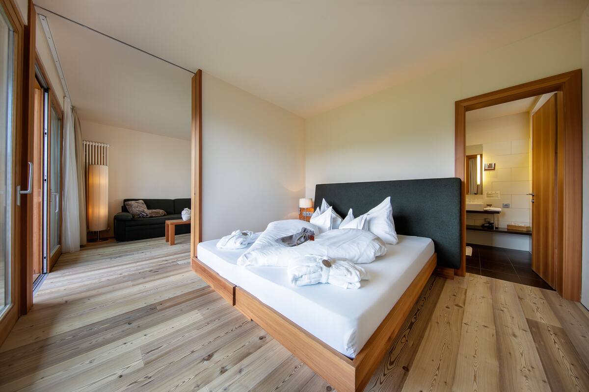 Habitación de hotel moderna con piso de madera, una cama grande con ropa blanca, cabecera oscura y vista al baño a través de una puerta abierta.