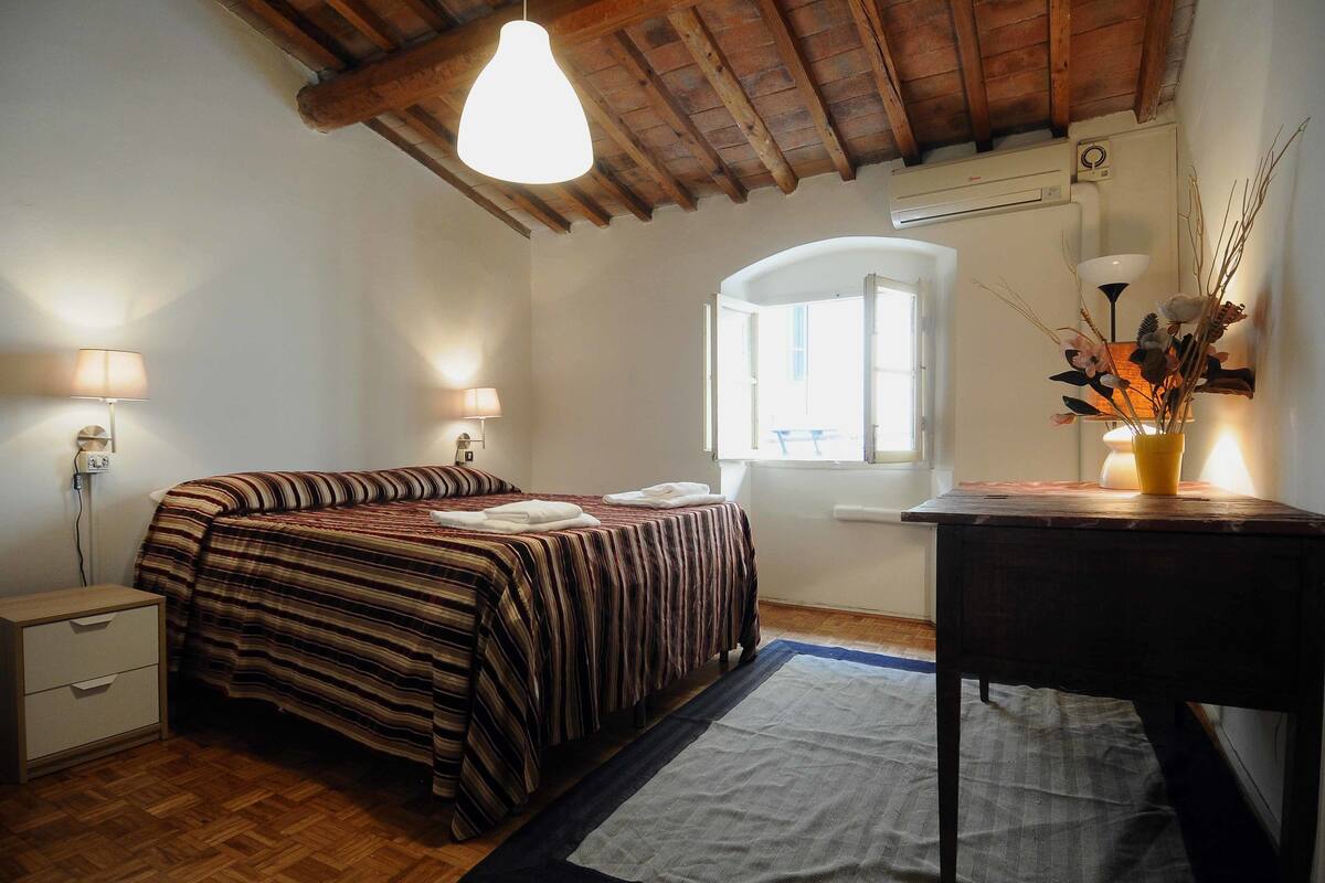A bedroom with a bed, bedside lamps, a wooden desk, a window, and a wooden ceiling.
