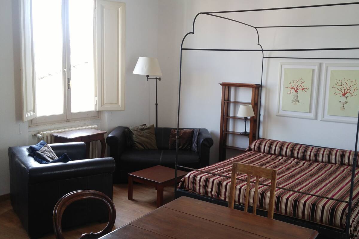 A room with a four-poster bed, a sofa, a small table, and a chair. There are two windows with curtains, a radiator, and wall-mounted lamps.