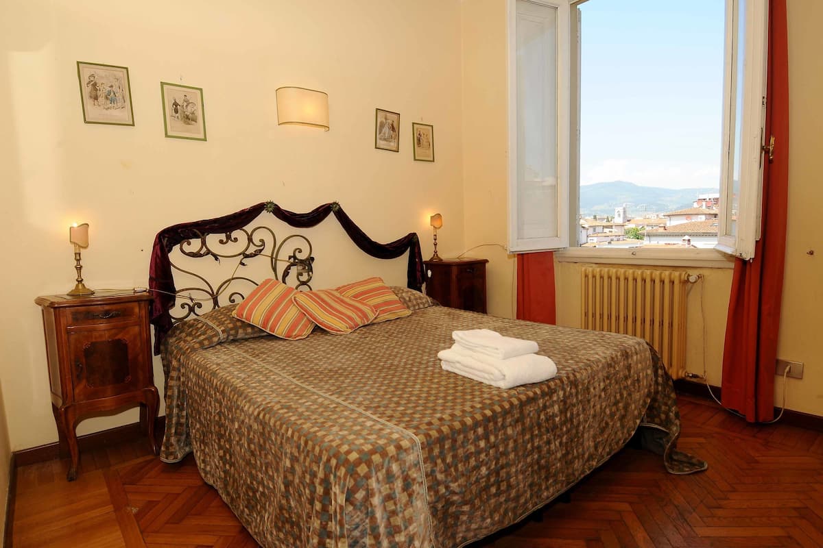 A neatly made bed with a patterned bedspread, two bedside lamps, and a window with a view of buildings and mountains.