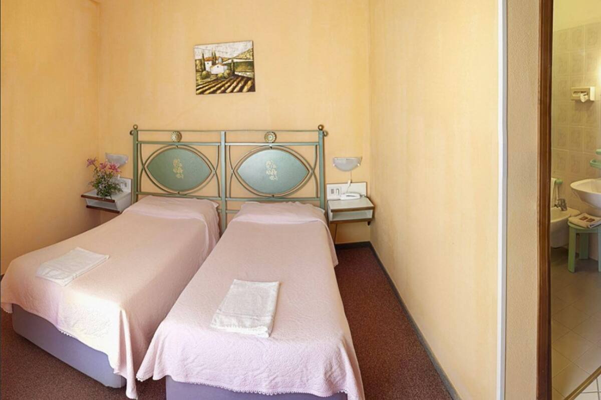 Ein Hotelzimmer mit zwei Betten, einem Nachttisch mit Lampe, einem kleinen Tisch mit Blumenvase und einem Bild an der Wand.