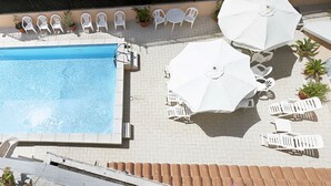 Een rechthoekig zwembad omringd door witte ligstoelen en parasols, met een betegeld terras en potplanten.