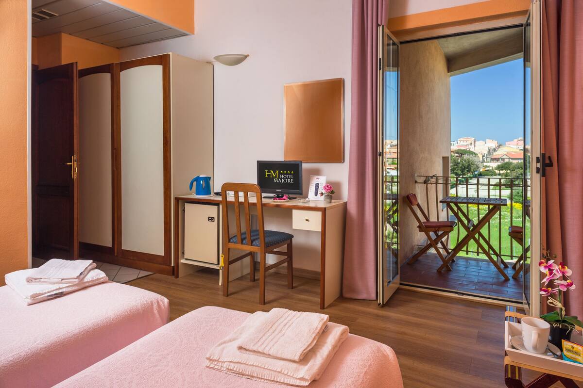 Ein Hotelzimmer mit zwei Betten, einem Schreibtisch mit Computer, einem Sessel und einem Balkon mit Blick auf die Stadt.