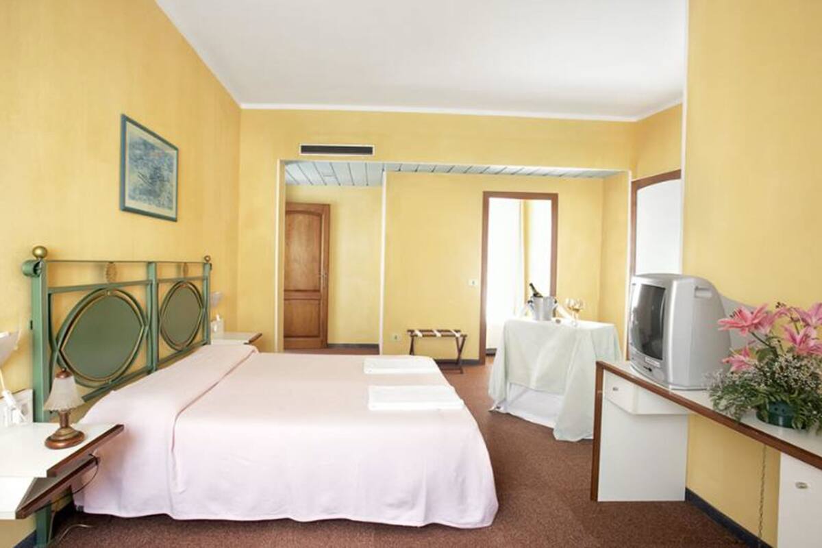 Ein Hotelzimmer mit einem Bett, einem Schreibtisch mit Fernseher, einem kleinen Tisch mit Flasche und Gläsern sowie einer Tür zu einem weiteren Raum.
