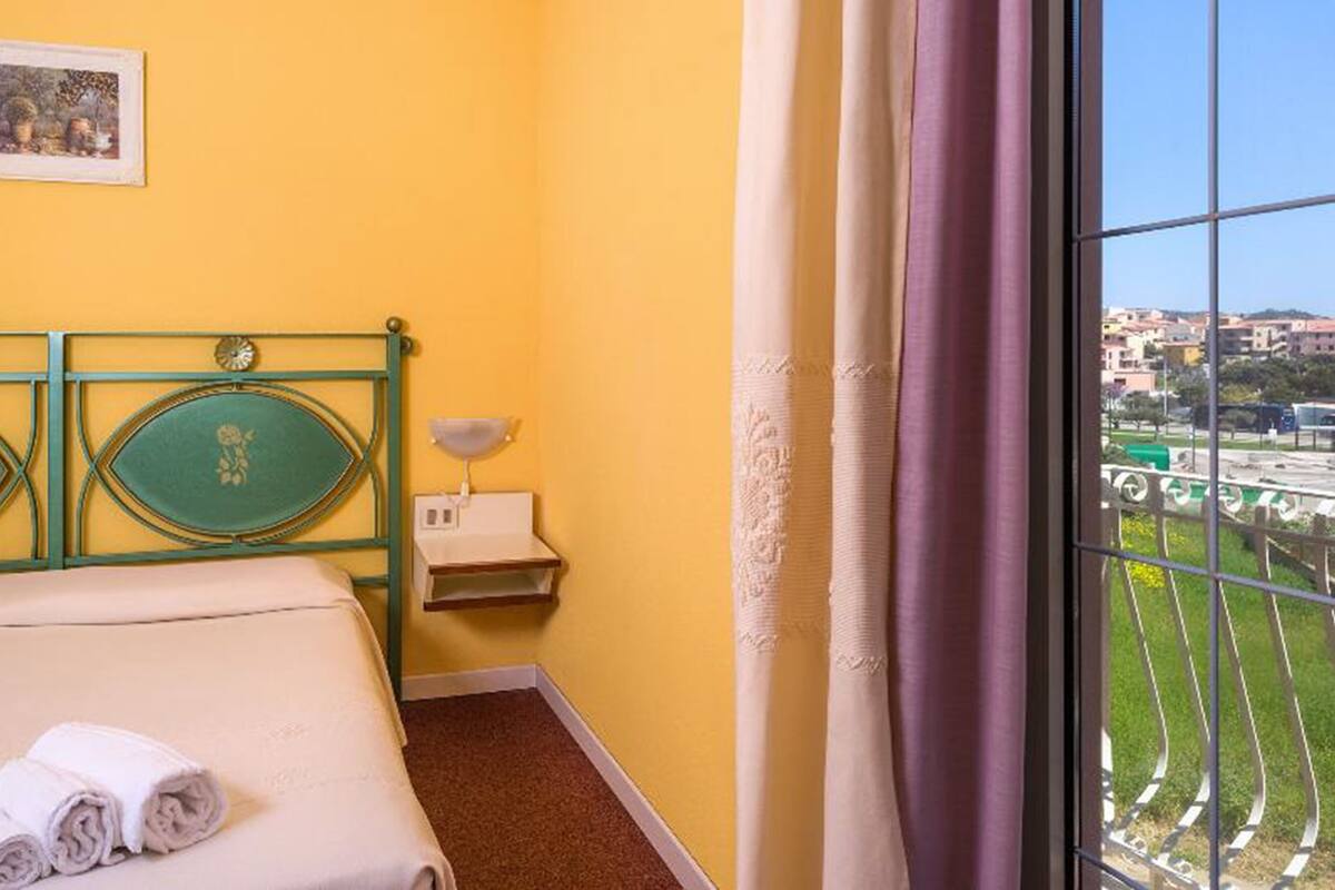 Ein Hotelzimmer mit Bett, Nachttisch, Lampe und einem Fenster mit Blick auf die Stadt.