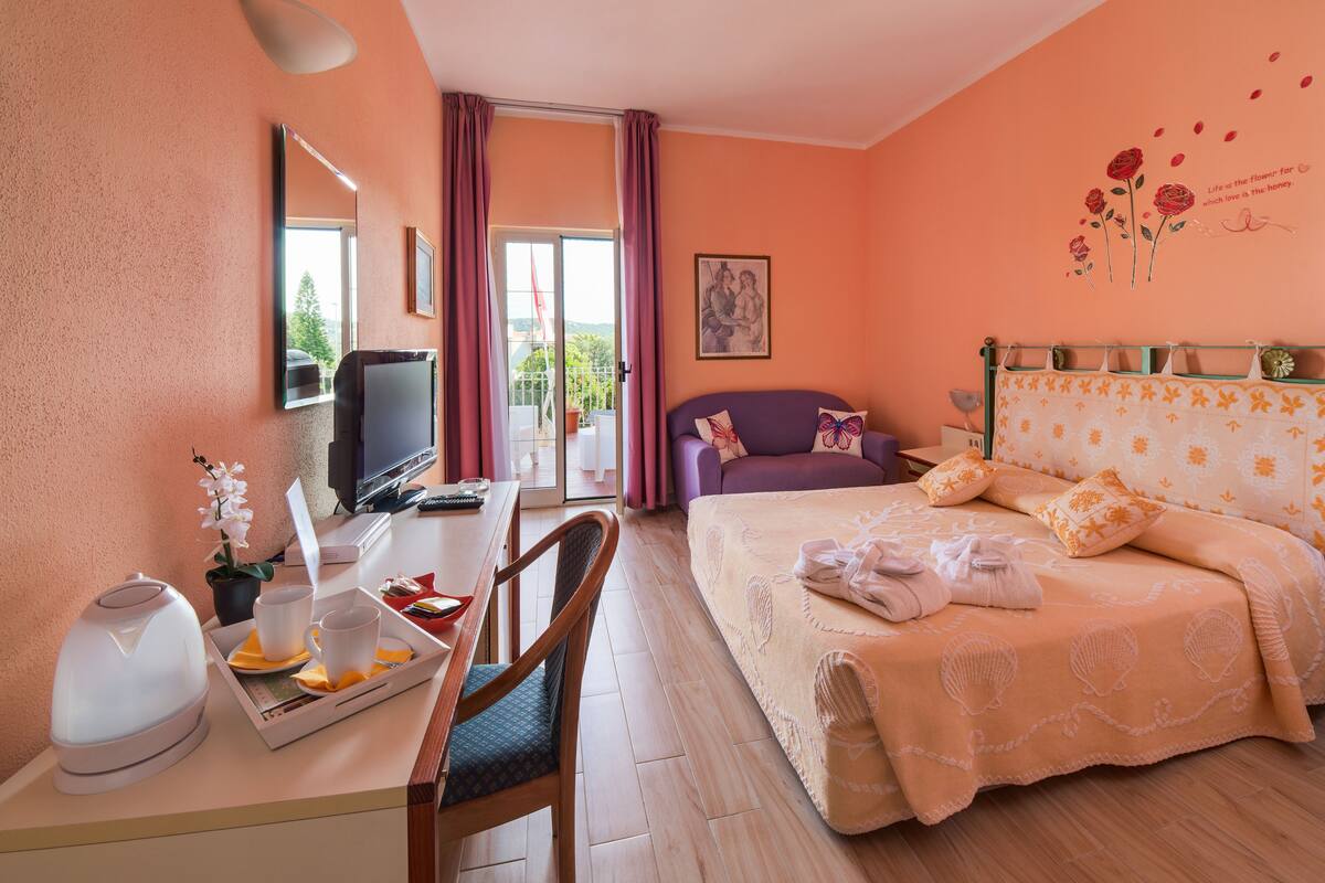 Ein Hotelzimmer mit einem Bett, einem Schreibtisch mit Wasserkocher, Tassen und Tablett, einem Sessel, einem Fernseher und einem Fenster mit Vorhängen.
