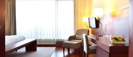 A hotel room with a bed, a desk, a chair, a lamp, and a large window with curtains.