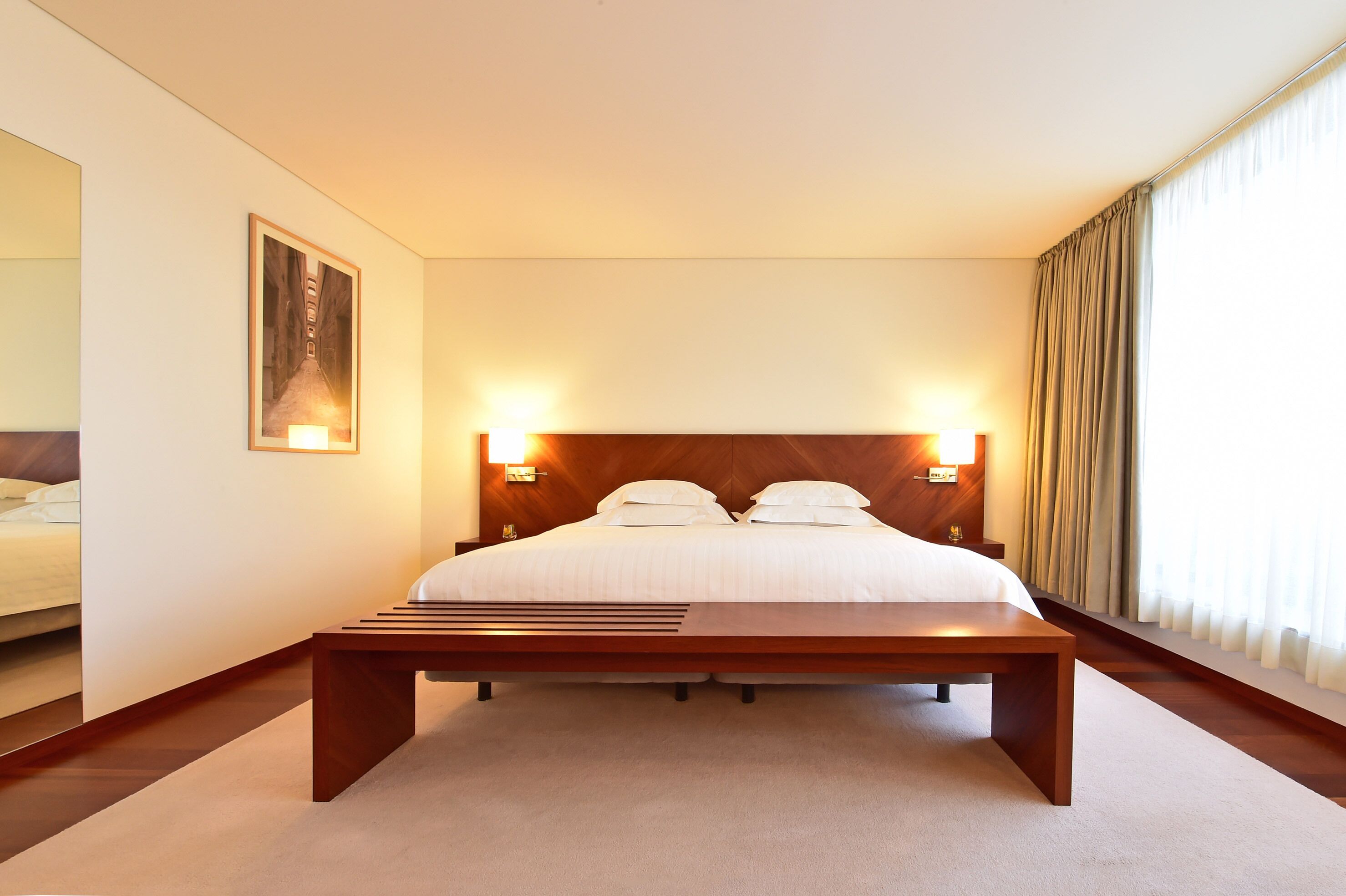 Habitación de hotel con una cama grande, lámparas de noche, cabecero de madera, un cuadro en la pared y una ventana con cortinas.