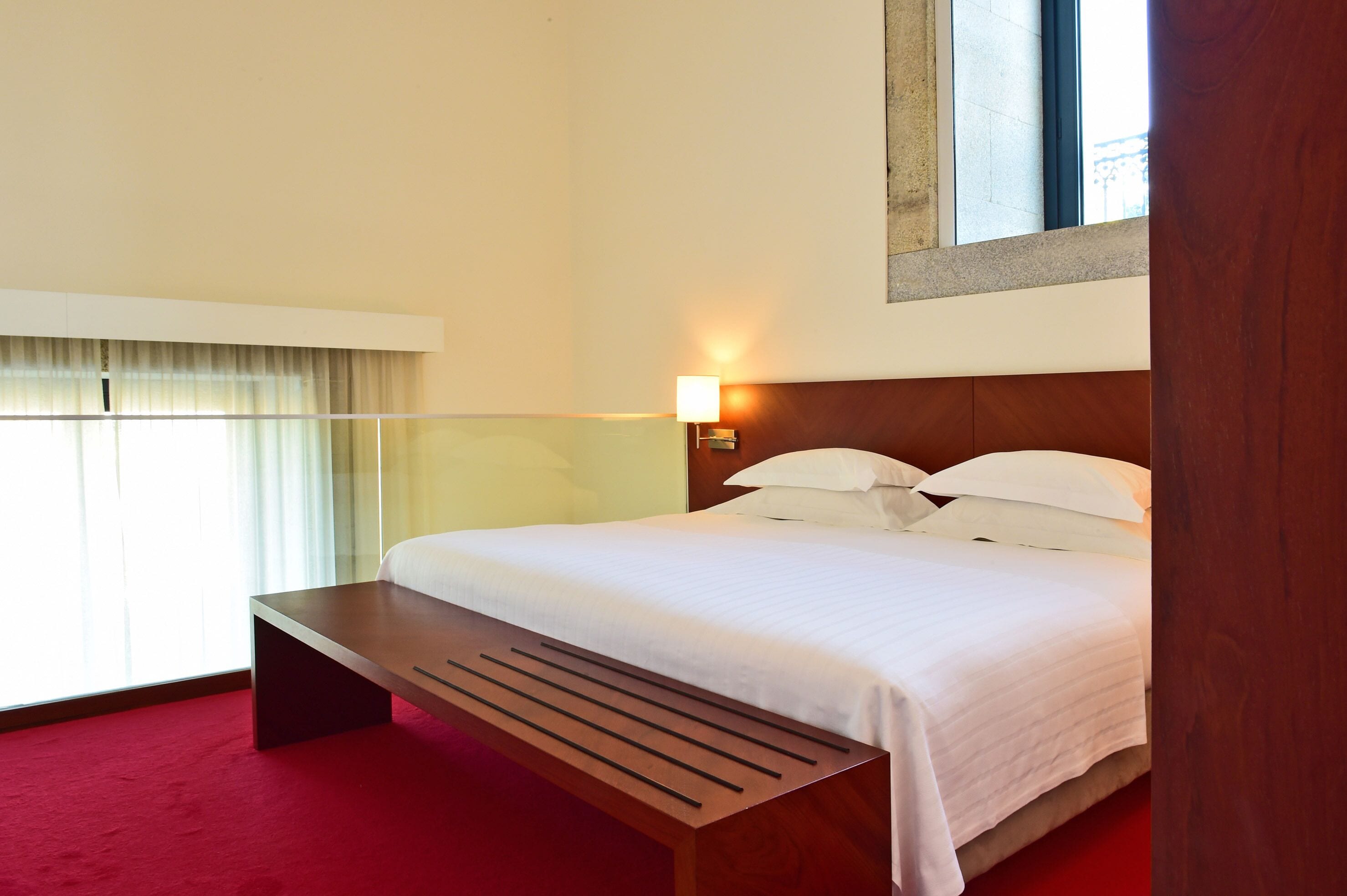 Una habitación de hotel con una cama, una mesita de noche, una lámpara y una ventana con cortinas.