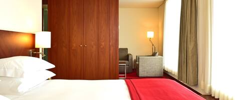 A hotel room with a large bed, a wooden wardrobe, a small armchair, and a desk with a lamp.