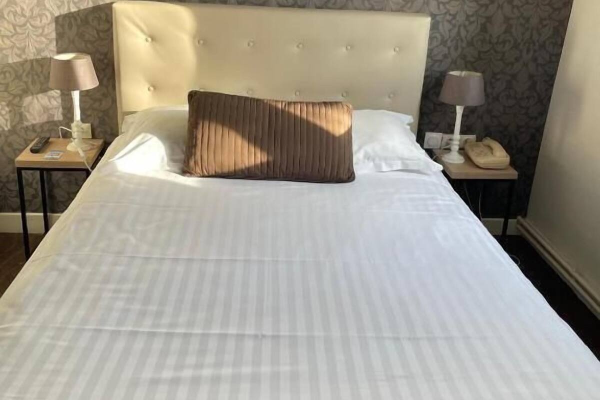 Un lit bien fait, recouvert d’une couvre-lit à rayures et agrémenté d’un seul oreiller marron, flanqué de deux tables de chevet sur lesquelles reposent des lampes et un téléphone.